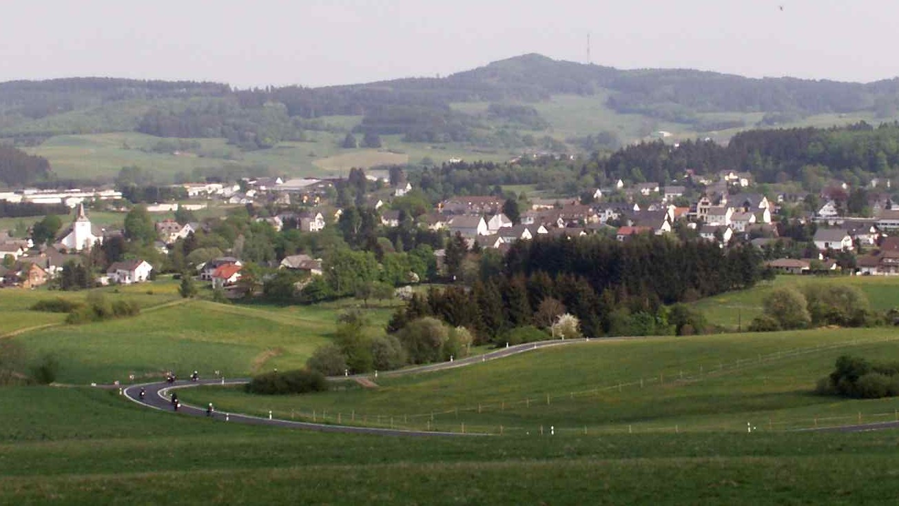 Geschwungene Landstraße durch die Wiesen bei Kelberg, dahinter der Ort und die Hohe Acht